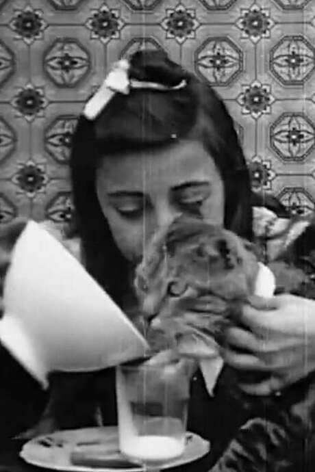 A young girl receives her breakfast of milk and a biscuit from an elderly woman. When her beribboned cat appears, she offers the food to the feline. The cat then dips its paw into the milk glass, licking it clean, creating a tender, everyday moment of shared comfort.